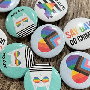 May include: A collection of ten 1-inch button badges with various designs. The badges feature a rainbow heart, a cat with rainbow sunglasses, a rainbow heart with a black and brown stripe, a rainbow heart with a blue and brown stripe, a rainbow heart with a pink and brown stripe, a rainbow heart with a white and brown stripe, a rainbow heart with a light blue and brown stripe, a rainbow heart with a dark blue and brown stripe, a rainbow heart with a purple and brown stripe, and a rainbow heart with a green and brown stripe. The badges are all white with black outlines.