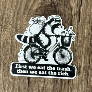 May include: Black and white sticker featuring a raccoon riding a bicycle, carrying a bag and basket of trash. The sticker has the text "First we eat the trash, then we eat the rich."