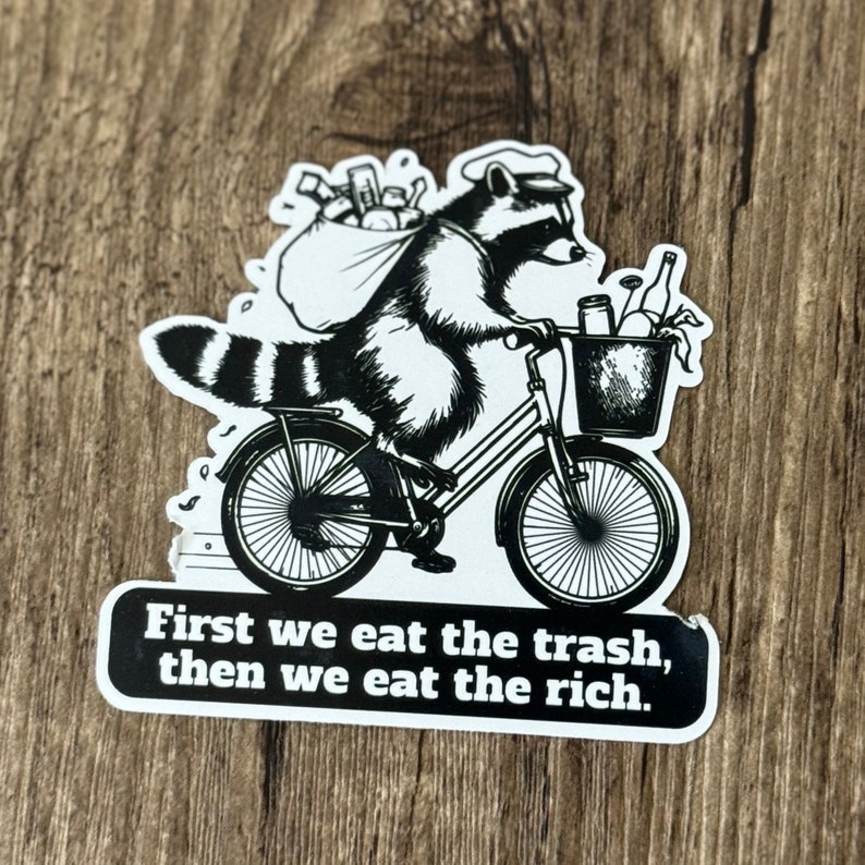 May include: Black and white sticker featuring a raccoon riding a bicycle, carrying a bag and basket of trash. The sticker has the text "First we eat the trash, then we eat the rich."