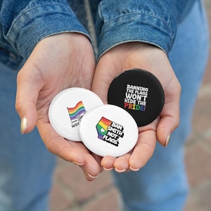 May include: Three circular badges with rainbow flag designs and text are held in open hands. One badge is black with white text: "Banning the flags won't hide the pride!" The other two are white with rainbow flag designs and text.