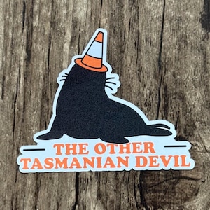 May include: A sticker featuring a black seal wearing an orange and white traffic cone on its head. Below the seal, the text reads THE OTHER TASMANIAN DEVIL in orange capital letters against a light blue background with a white border.