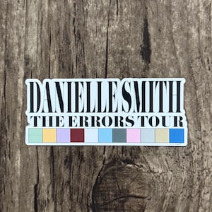 May include: A rectangular sticker with the text "DANIELLE SMITH THE ERRORS TOUR" in a bold, stylized font. The sticker has a white background with a colorful stripe of squares at the bottom. The sticker is placed on a wooden surface.