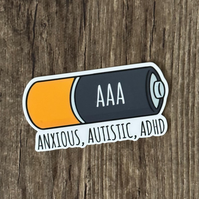 K&ouml;nnte beinhalten: Wei&szlig;er Aufkleber mit einer Cartoon-Zeichnung einer AAA-Batterie. Die Batterie ist orange und schwarz. Der Text "ANXIOUS, AUTISTIC, ADHD" steht unter der Batterie.