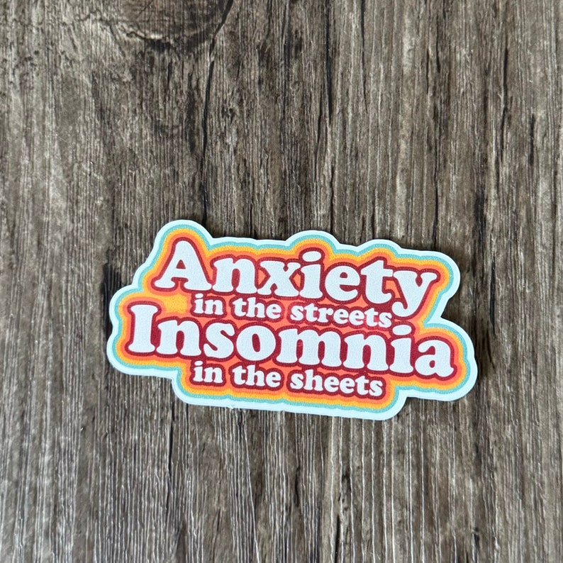 以下が含まれることがあります： 虹色の縁取りのあるレトロなフォントで「Anxiety in the streets Insomnia in the sheets」と書かれたステッカーです。