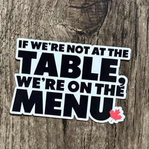 May include: A white sticker with black text that reads "IF WE'RE NOT AT THE TABLE WE'RE ON THE MENU" with a red maple leaf. The sticker is on a wooden surface.