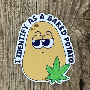 May include: A sticker with a cartoon potato with a relaxed expression and the text "I IDENTIFY AS A BAKED POTATO." A green cannabis leaf is at the bottom. The sticker is on a wooden surface.