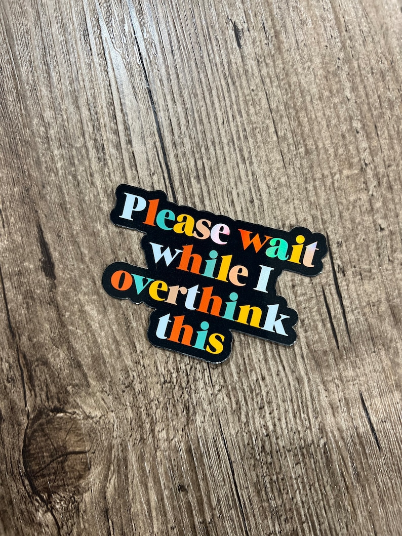 May include: A black sticker with the text "Please wait while I overthink this" in colorful letters. The sticker is on a wood background.
