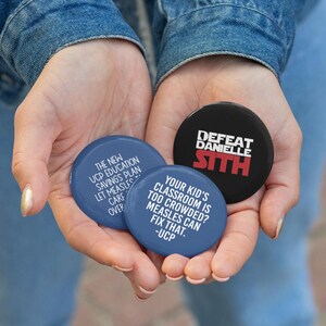 May include: Three round buttons with text are held in open hands. Two blue buttons read "THE NEW UCP EDUCATION SAVINGS PLAN LET MEASLES CARE OVER" and "YOUR KID'S CLASSROOM IS TOO CROWDED? MEASLES CAN FIX THAT. -UCP". The third button is black and says "DEFEAT DANIELLE SITH".