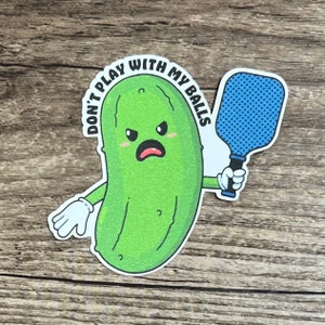May include: A green pickle with a cartoon face holding a blue paddleball racket. The pickle is wearing white gloves and has the text "Don't play with my balls" written around it.