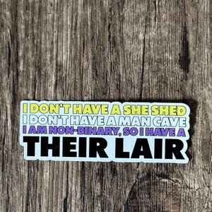 May include: A rectangular sticker with the text "I DON'T HAVE A SHE SHED I DON'T HAVE A MAN CAVE I AM NON-BINARY, SO I HAVE A THEIR LAIR" in yellow, purple, and black lettering. The sticker is on a wooden surface.