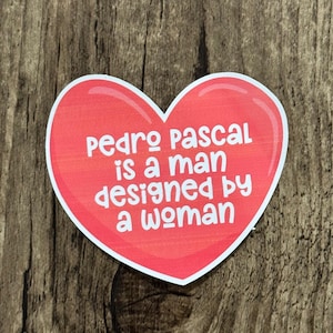 May include: A heart-shaped sticker with a red background and white border. The sticker features the text "pedro pascal is a man designed by a woman" in white, sans-serif font. The sticker is placed on a wooden surface.