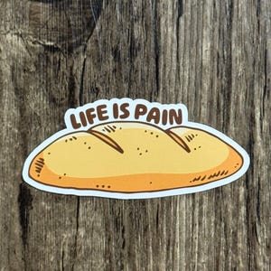 Peut inclure: Un autocollant représentant une miche de pain de dessin animé avec le texte "LIFE IS PAIN" en marron. Le pain est jaune et orange avec des détails bruns, sur un fond en bois.