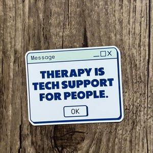 Peut inclure: Un autocollant blanc avec une bordure bleue et le texte "THERAPY IS TECH SUPPORT FOR PEOPLE." L'autocollant a un en-tête bleu clair qui dit "Message" et un petit bouton "OK". L'autocollant est sur une surface en bois.