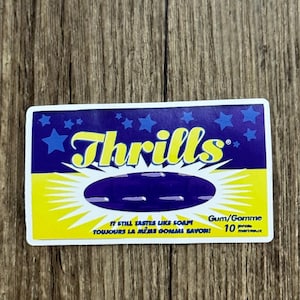 May include: A rectangular sticker with the word "Thrills" in yellow script on a dark blue background with blue stars. The sticker also includes the text "IT STILL TASTES LIKE SOAP! TOUJOURS LA MÊME GOMME SAVON!" and "Gum/Gomme 10 pcs".
