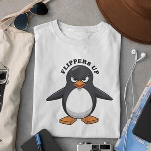 Könnte beinhalten: Weißes T-Shirt mit einem Cartoon-Pinguin, der einen schwarz-weißen Smoking trägt. Der Pinguin sieht wütend aus und der Text "FLIPPERS UP" ist in Schwarz über dem Pinguin geschrieben.