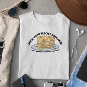 Puede incluir: Una camiseta blanca de manga corta con un gráfico de una pila de tortitas con sirope y mantequilla en un plato azul. El texto dice FUCK YOUR FASCIST PANCAKES y I DON'T WANT YOUR SEPARATIST STACK, DANI en letras mayúsculas negras.