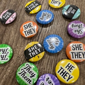May include: A collection of colourful round buttons with various text, including "She They", "He They", "They Them", and "She They". The buttons are on a wooden surface.