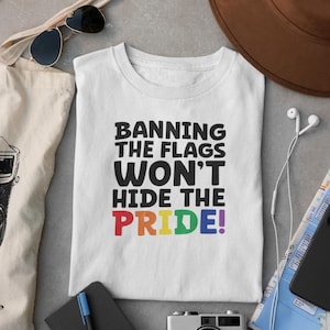 May include: White t-shirt with the text "Banning the Flags Won't Hide the Pride!" in black and rainbow colors. The shirt is folded and lies on a gray surface, with sunglasses, a tote bag, and a hat visible.