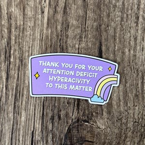 May include: A purple sticker with the text "THANK YOU FOR YOUR ATTENTION DEFICIT HYPERACTIVITY TO THIS MATTER." The sticker has a rainbow and cloud graphic. The sticker is on a wooden surface.