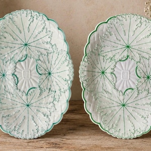 Puede incluir: Dos platos ovalados de cerámica con base blanca, patrones de hojas en relieve y contornos pintados en verde. Los platos tienen un borde festoneado y un motivo cuadrado central. El diseño incluye detalles intrincados de las nervaduras.