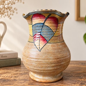 Peut inclure: Un vase en céramique au corps beige texturé avec des stries horizontales. La partie supérieure présente un motif de feuille peint à la main dans des tons rouge, bleu et jaune, avec une ouverture festonnée à bord sombre et une base large.