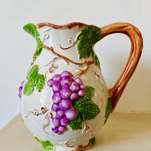 May include: White ceramic pitcher with a brown handle and grape cluster design. The pitcher features a raised relief of purple grapes, green leaves, and brown vines. The handle is a curved, solid piece. A decorative piece for serving.