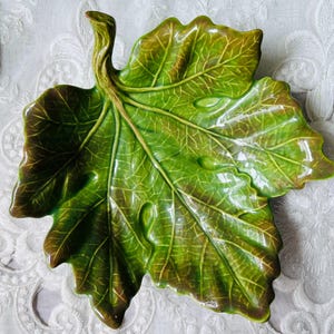 May include: A ceramic dish shaped like a large grape leaf with a glossy finish. The surface features a vibrant green color with deep brown shading along the edges and veins, mimicking the natural appearance of a leaf with a prominent stem.