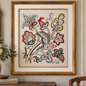 May include: A framed embroidery piece featuring a central floral motif with intricate stitching in shades of red, blue, gold, and green on a light beige fabric background. The artwork is encased in a rectangular gold frame with a white mat border.
