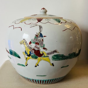 May include: A white ceramic jar with a lid, featuring a hand-painted scene of a figure on a yellow horse. The jar has a rounded shape with a decorative rim and lid. The artwork includes green, red, and blue accents.