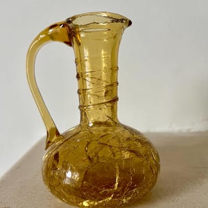 Puede incluir: Una pequeña jarra de vidrio ámbar con un acabado craquelado y un asa. La jarra mide aproximadamente 15 cm de alto.