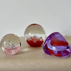 Puede incluir: Tres pisapapeles de vidrio con remolinos rojos y blancos en su interior. Un pisapapeles es morado, otro es transparente con una base rosa y el último es transparente con una base roja.