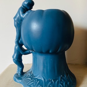 May include: A blue ceramic vase with a unique design. Two figures are climbing the side of the vase. The vase has a bulbous top and a flared base with a grass and flower design. The vase is approximately 7 inches tall.
