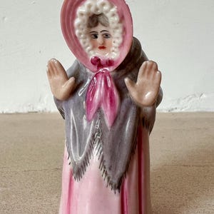 Könnte beinhalten: Vintage Keramikfigur einer Frau in rosa Haube und Kleid, mit grauem Schal und offenen Händen. Die Figur hat ein detailliertes Gesicht und ist etwa 15 cm hoch.