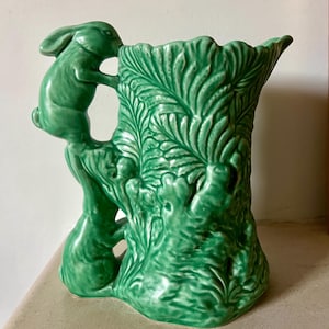 Może przedstawiać: Zielony ceramiczny dzban z królikiem wspinającym się po boku. Dzban ma kształt pnia drzewa ze szczegółowymi wzorami liści. Królik jest umieszczony blisko krawędzi, dodając fantazyjnego akcentu do tego dekoracyjnego elementu.