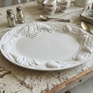 May include: White oval serving platter with embossed lobster and seashell designs. The rim features a repeating pattern of waves, shells, and a lobster. The platter is on a distressed white wooden table, with salt and pepper shakers and teacups in the background.