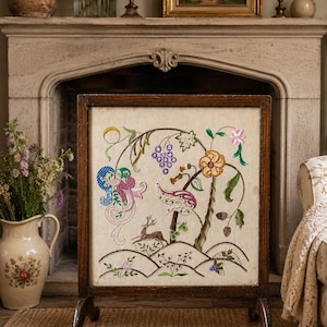 May include: A rectangular fireplace screen with a dark wood frame and a cream-colored fabric panel. The fabric features colorful embroidery depicting a deer, grape clusters, floral motifs, and stylized vines in shades of pink, purple, yellow, and green.