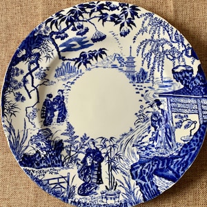Könnte beinhalten: Ein weißer Keramikteller mit einem japanisch inspirierten Design in Blau und Weiß. Der Teller zeigt eine Landschaftsszene mit einer Pagode, Bäumen und Figuren in traditioneller Kleidung.