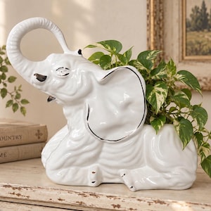 Pode incluir: Um vaso de cerâmica branca em forma de elefante com acabamento brilhante, apresentando uma tromba levantada e detalhes pintados a preto nos olhos e presas. O dorso oco do elefante contém uma planta pothos variegada verde e branca com folhas.