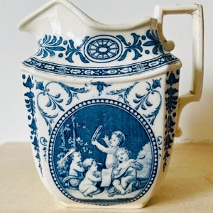 May include: Cream-colored ceramic pitcher with blue floral and cherub design. The pitcher features a handle and spout, with a detailed blue illustration of cherubs playing musical instruments. The design is repeated around the top and sides of the pitcher.