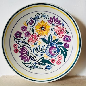 Puede incluir: Un plato de cerámica blanco con un diseño floral colorido. El plato presenta una disposición central de flores en tonos amarillos, rosas, morados y azules, rodeadas de hojas verdes. El borde tiene un borde amarillo y verde.