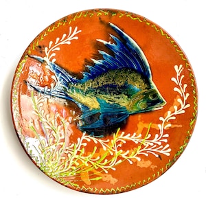 May include: A round ceramic plate with a glossy orange background featuring a central blue and green fish with prominent fins. The plate is decorated with white seaweed-like patterns and a green stitched-style border around the outer edge.