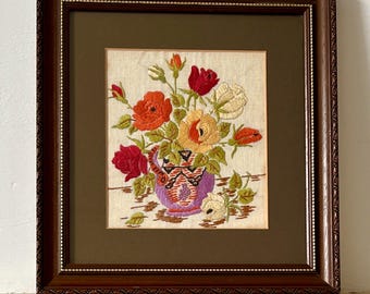 Op de afbeelding: Geborduurd bloemenkunstwerk in een donkerhouten lijst. Het kunstwerk toont een vaas gevuld met rode, oranje, gele en witte bloemen. De vaas is paars met een patroon. De lijst heeft een decoratieve rand en een donkergroene passe-partout.