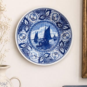 May include: A round ceramic decorative plate featuring a blue and white transferware pattern. The centre depicts a nautical scene with sailboats on water and a distant windmill. The border is decorated with intricate floral and leaf motifs.