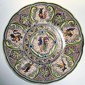 Puede incluir: Un plato redondo de cerámica con borde festoneado, que presenta un gallo pintado a mano en el centro, rodeado de intrincados patrones florales y de volutas en tonos verde, azul, naranja y marrón sobre un fondo color crema.