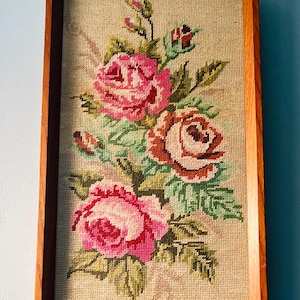 May include: A framed needlepoint featuring a bouquet of pink and red roses with green leaves. The artwork is set against a beige background and framed in a dark wood. The floral design is a classic decorative piece.