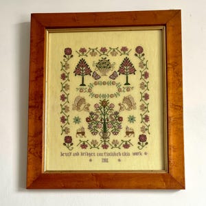 May include: Framed cross-stitch embroidery with a light yellow background. The design features trees, flowers, squirrels, and deer in shades of pink, green, and brown. The text reads "betty and bridget cox finished this work * 1981 *."