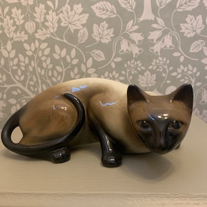 Può includere: Una statuetta di ceramica di un gatto siamese con uno schema di colori marrone e nero. Il gatto è sdraiato con la testa girata di lato e gli occhi aperti. La statuetta è realizzata in ceramica smaltata e ha un aspetto realistico.