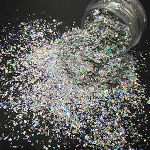 May include: A close-up of iridescent silver glitter, scattered on a black surface. The glitter is in the shape of small, irregular pieces.