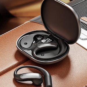 May include: A black and silver wireless earbud set with a charging case. The case is open, revealing the earbuds inside, and has a digital display. One earbud is separate, showing the design. The set is on a brown leather surface.