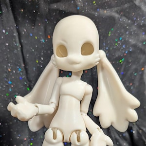May include: A cream-colored articulated doll with a stylized, rounded head, large eye sockets, and long, wing-like appendages. The doll has jointed limbs and is posed against a dark, glittery background. The doll's feet have a paw-like appearance.